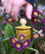 A child reaching for the lid of the Grapat yellow Little Things that has been placed amongst flowers outdoors. A wooden top with eyes drawn on and a yellow coloured pot to match. Grapat have a wide range of wooden toys and loose parts available at Babipur. 