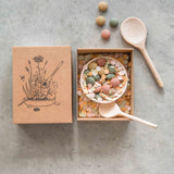 Grapat Yummy Wooden Play Food Set. The set comes with two wooden spoons a bowl and coloured pebbles in pastel shades. Pictured with the cardboard gift box that it comes in.