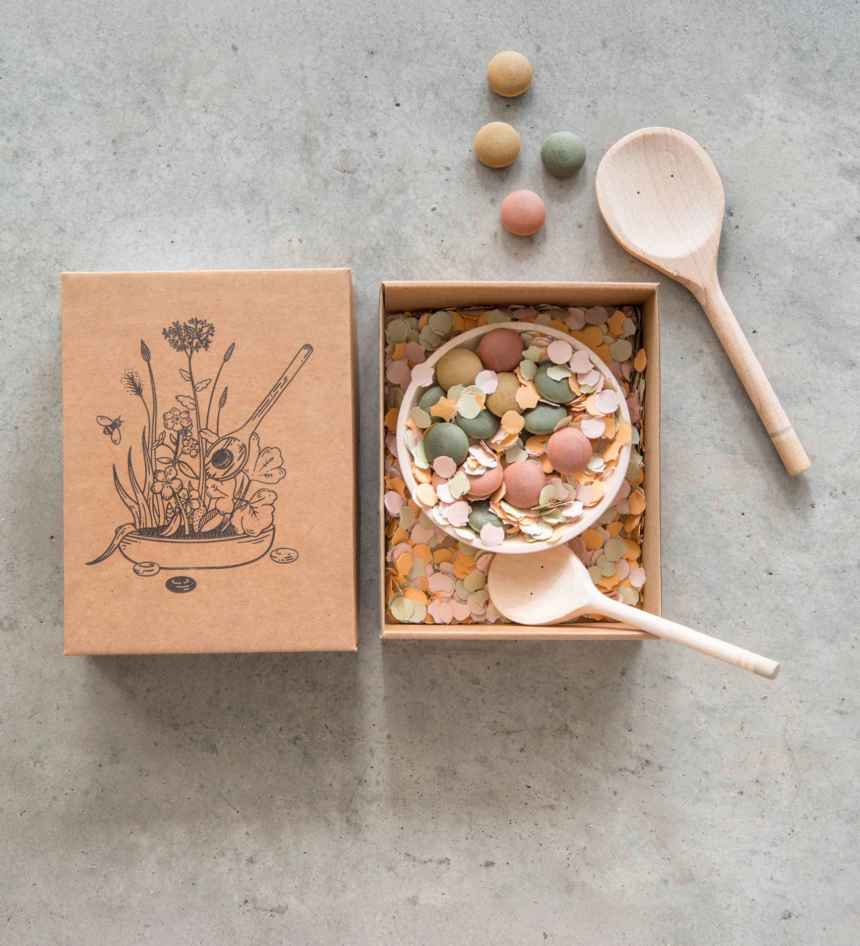 Grapat Yummy Wooden Play Food Set. The set comes with two wooden spoons a bowl and coloured pebbles in pastel shades. Pictured with the cardboard gift box that it comes in.