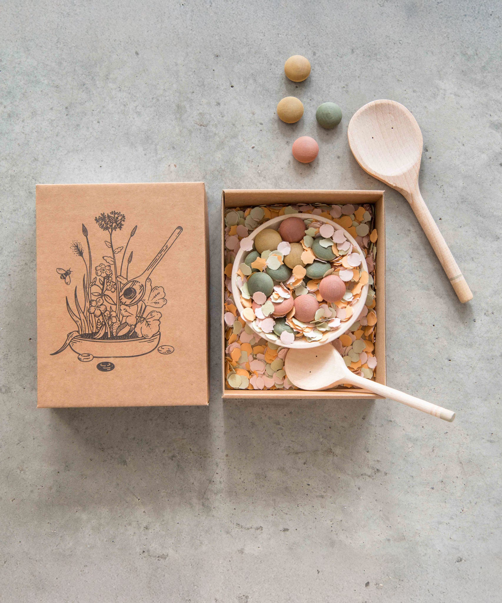 Grapat Yummy Wooden Play Food Set. The set comes with two wooden spoons a bowl and coloured pebbles in pastel shades. Pictured with the cardboard gift box that it comes in.