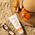 Green People SPF15 Edelweiss Tan Accelerator Sun Lotion placed on the sand next to a straw hat and a pair of sunglasses.