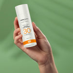 A hand holding up a tube Green People Facial Sun Cream SPF30 50ml against a green background. 