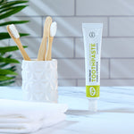 Green People Fennel & Propolis Natural Toothpaste on a marble surface in a bathroom next to a ceramic pot with bamboo toothbrushes.