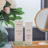 A Green People Gentle Cleanse & Make-up Remover 150ml next to the cardboard box packaging on wooden bamboo tray. There is a round bathroom mirror next to them and a vase of flowers on teh other side.