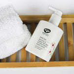 A bottle of Green People Manuka & Lemon Tea Tree Hand Wash placed on a bamboo bath shelf next to a fluffy white towel. 