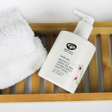 A bottle of Green People Manuka & Lemon Tea Tree Hand Wash placed on a bamboo bath shelf next to a fluffy white towel. 