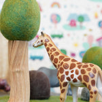 Close up of a green rubber toys giraffe figure on some green felt next to a papoose wooden tree toy