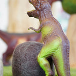 Close up of a green rubber toys t-rex stood on some green felt in front of a papoose felt rock toy