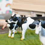 Close up of the green rubber toys cow figures stood on some green felt in front of a wooden house toy