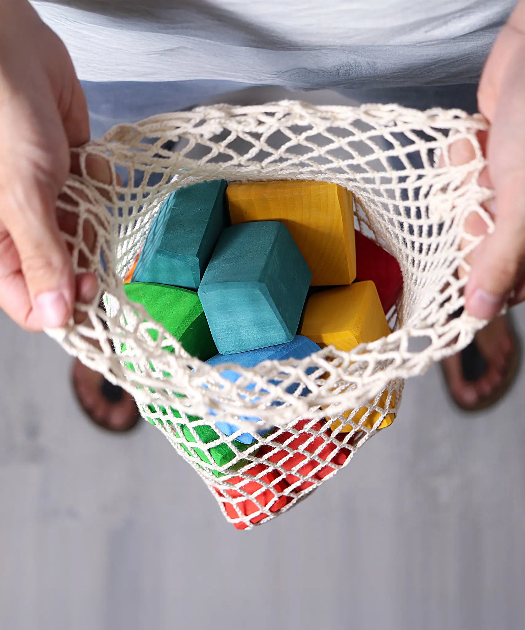 Adults holding the Grimm's 15 rainbow coloured wooden toy building blocks in the net storage bag.