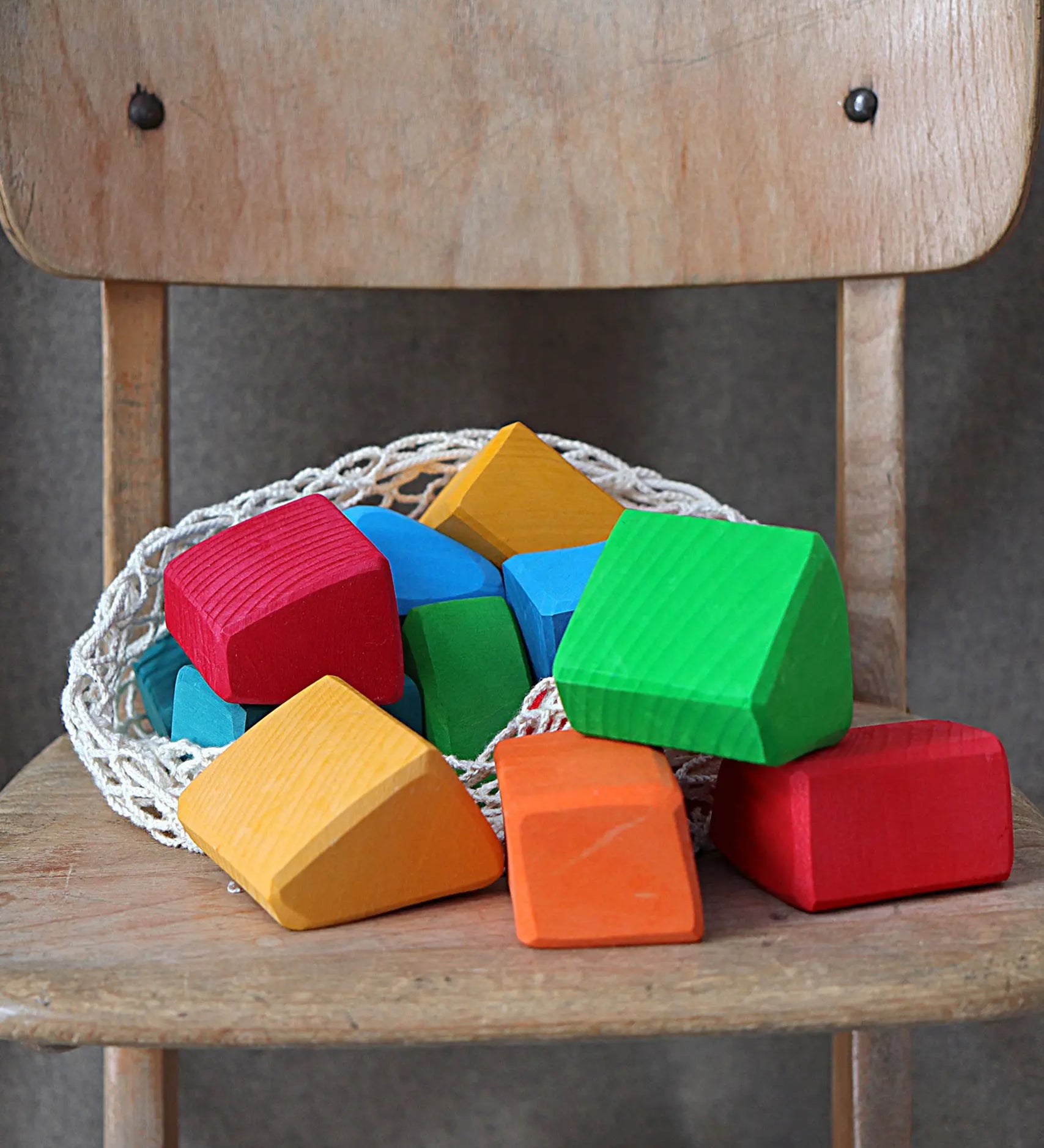 Grimm's 15 rainbow coloured wooden toy building blocks in a net storage bag on a wooden chair.