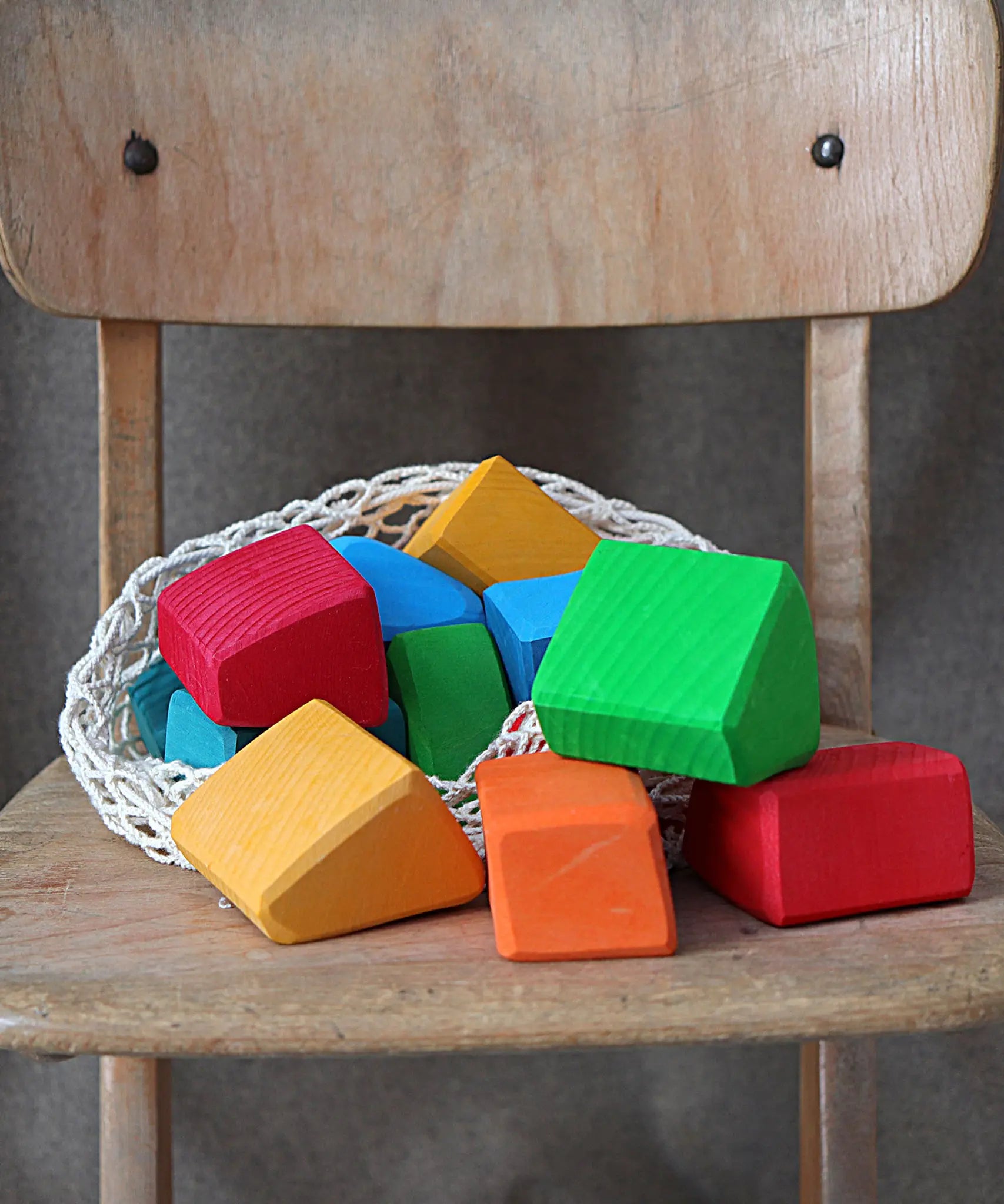 Grimm's 15 rainbow coloured wooden toy building blocks in a net storage bag on a wooden chair.