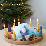 A Grimm's 16-Hole Natural Celebration Ring decorated in a Winter theme with candles, Nins, Magos, Grimm's dwarfs, pine cones. A Christmas tree with twinkling fairy lights can be seen in the background. 