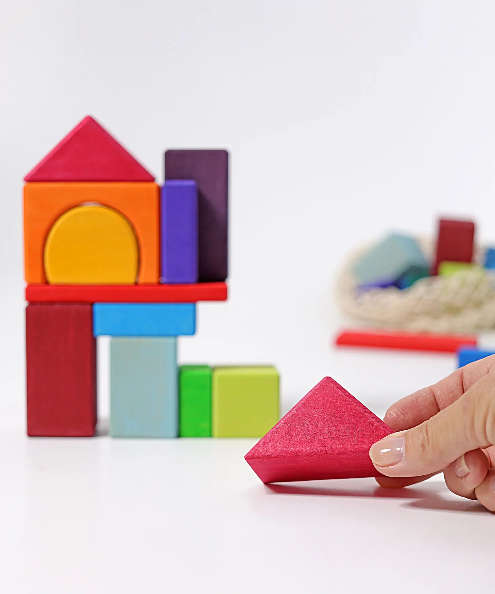 Grimm's 30 Coloured wooden Geo Blocks, an adult's hand can be seen holding one of the triangle shaped pieces.