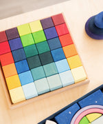 Grimm's 36 rainbow cubes wooden blocks in a storage tray next to a polar light set and purple wooden pebbles