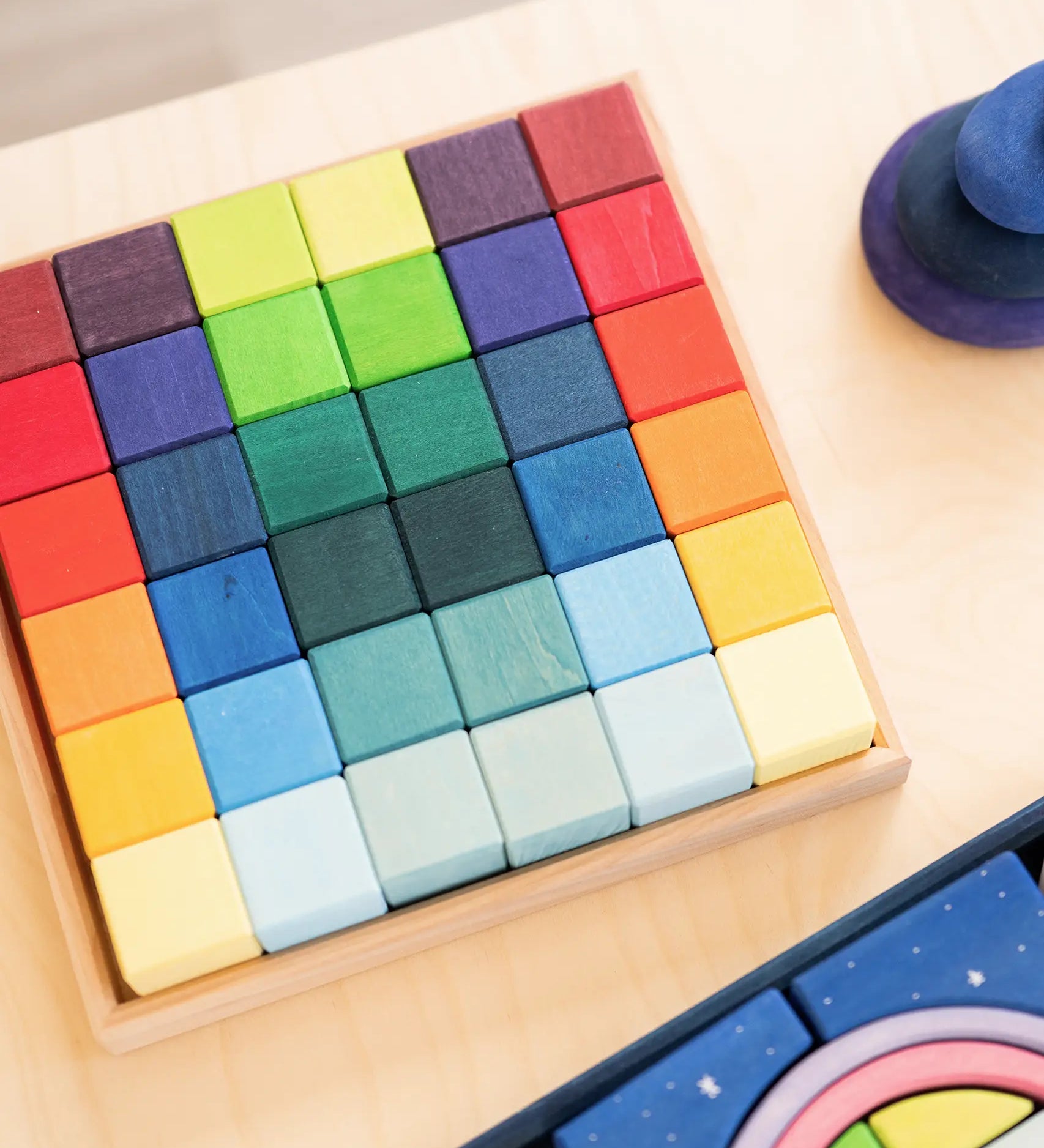 Grimm's 36 rainbow cubes wooden blocks in a storage tray next to a polar light set and purple wooden pebbles