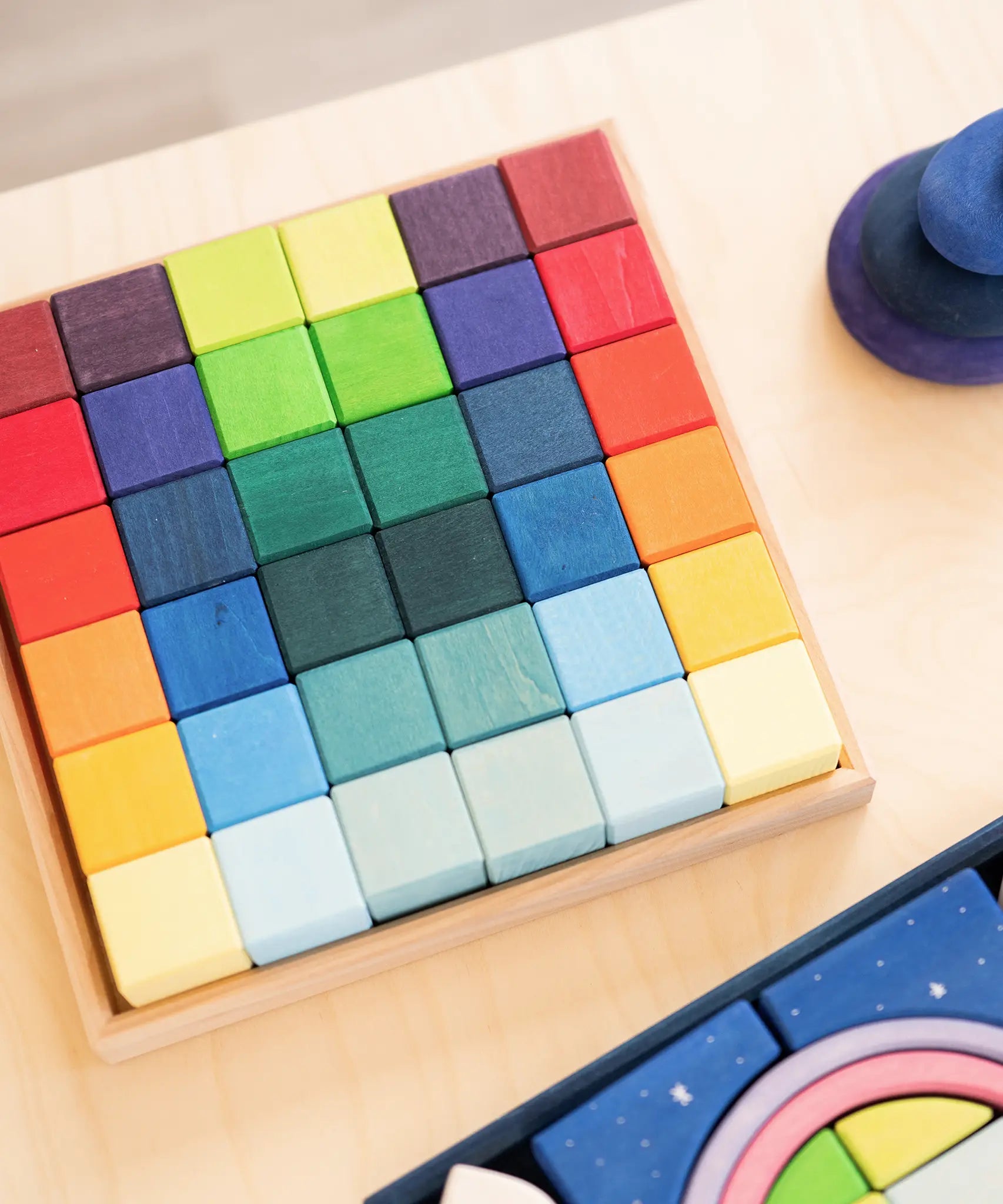 Grimm's 36 rainbow cubes wooden blocks in a storage tray next to a polar light set and purple wooden pebbles