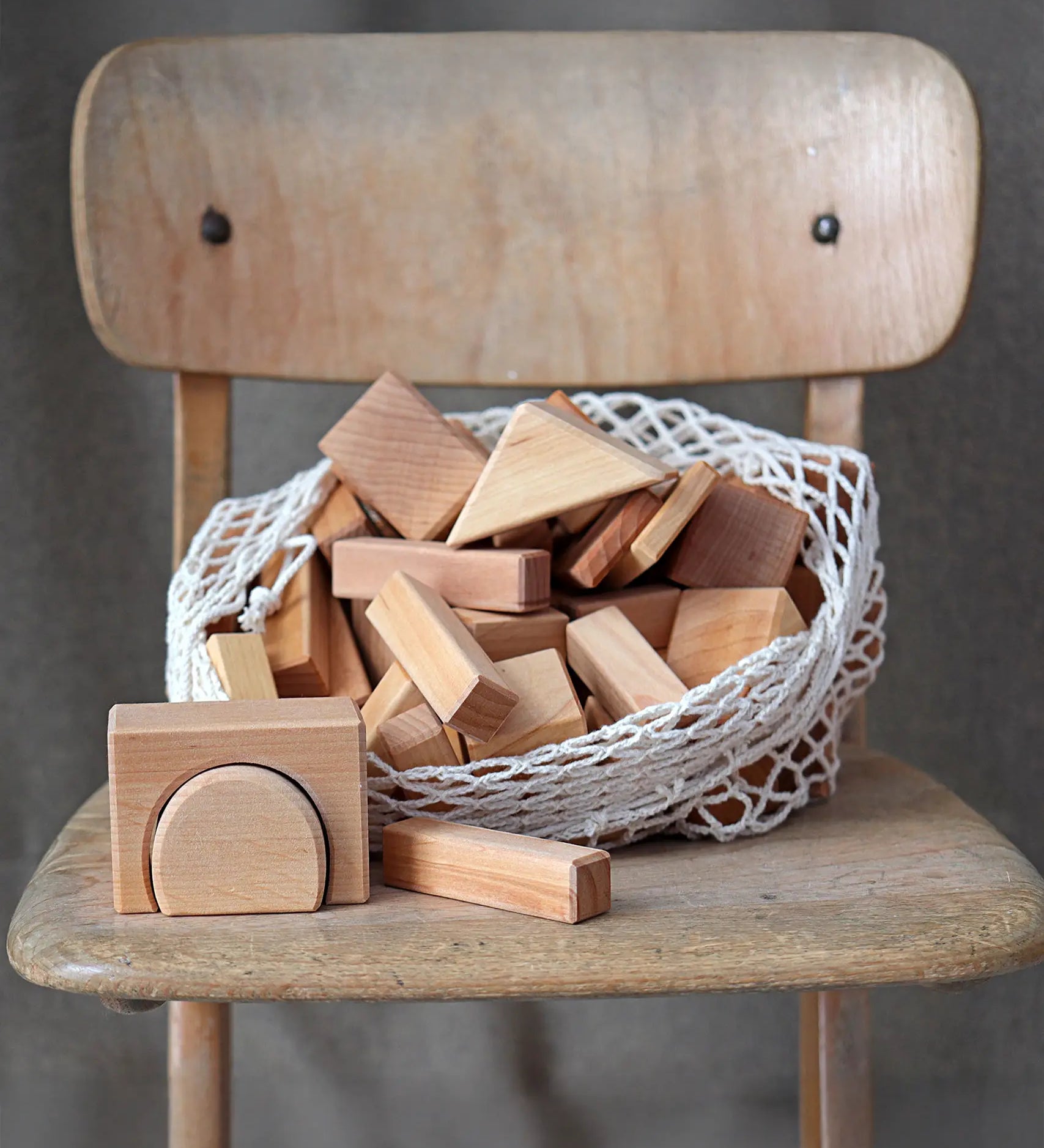 Grimm's 60 natural geo wooden building blocks in a net storage bag placed on a wooden chair.
