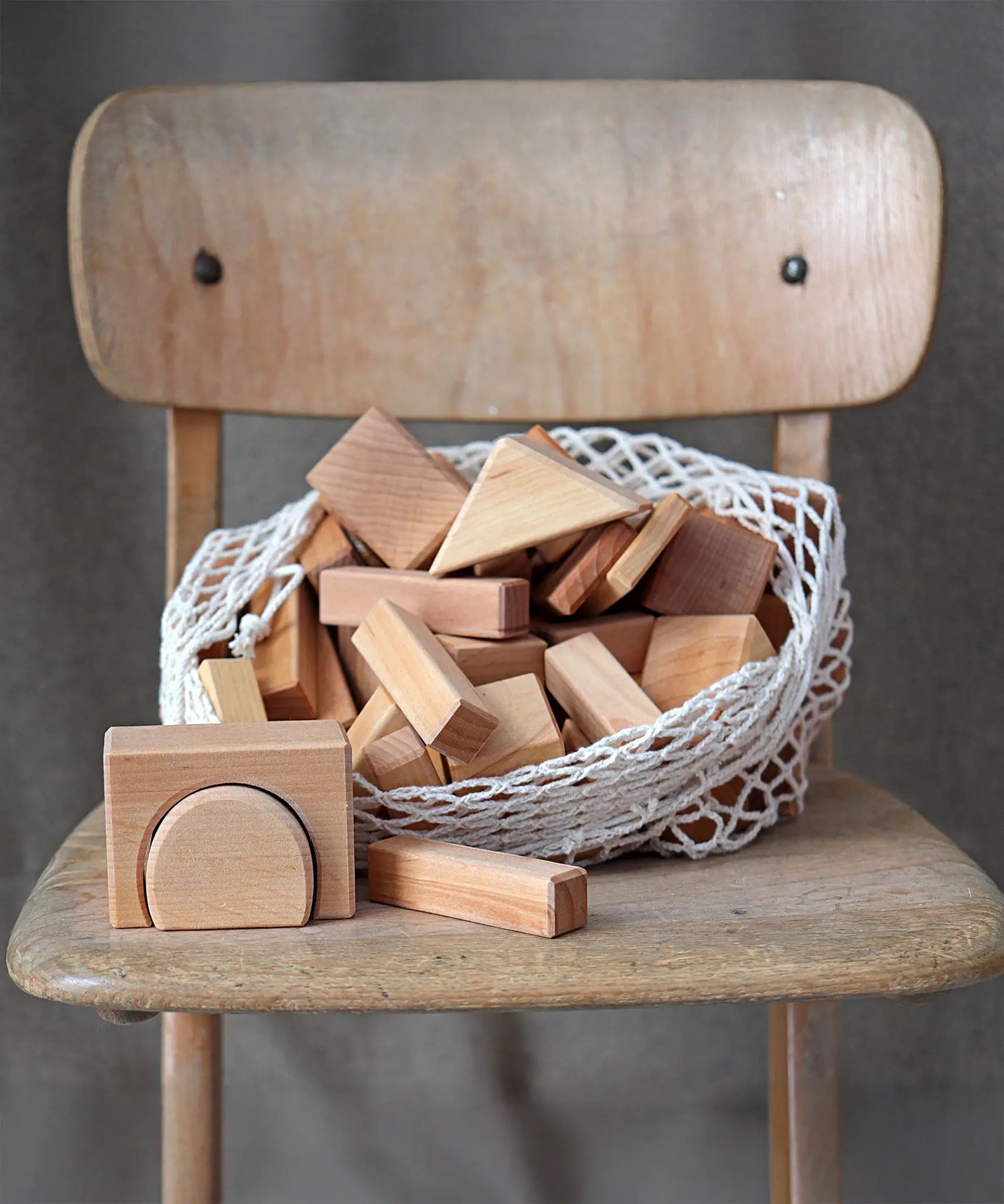 Grimm's 60 natural geo wooden building blocks in a net storage bag placed on a wooden chair.