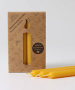 A pack of 12 Grimm's amber 100% beeswax candles in a cardboard box with three candles at the front laid out in front of the box.