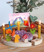 A Autumn display with the Grimms celebration ring with different wooden decorative figures on top including a kite, windmill, flowers, Autumn dwarf, mushroom and beeswax candles