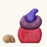 Grimm's purple bakery block house wooden toy next to a snail shell. 