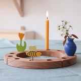 A Grimm's bee decorative figure placed on a natural Grimm's celebration ring. There is a lit candle, yellow tulip and a blue ceramic vase also placed on the ring. The bee is part of a wide range of Waldorf inspired decorative figures available here at Babipur. 