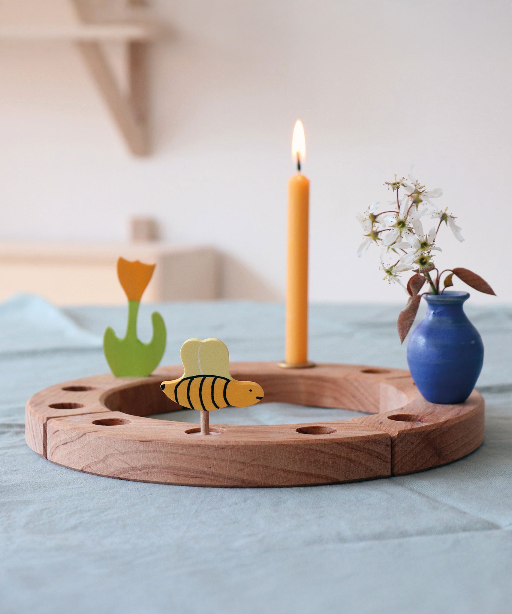 A Grimm's bee decorative figure placed on a natural Grimm's celebration ring. There is a lit candle, yellow tulip and a blue ceramic vase also placed on the ring. The bee is part of a wide range of Waldorf inspired decorative figures available here at Babipur. 