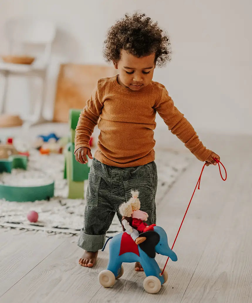 Child pulling the Grimms handcrafted wooden blue elephant in a playroom with a waldorf doll sitting on top 