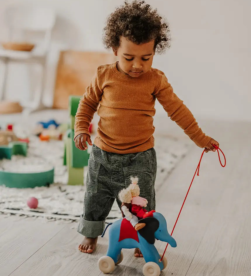 Child pulling the Grimms handcrafted wooden blue elephant in a playroom with a waldorf doll sitting on top 