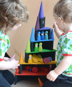 Children sitting next o a stack made from Grimms bright rainbow building blocks including semi circles and arch pieces