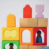The blocks from the Grimm's Windows Building Set wooden blocks placed in a stack on top of each other. Grimm's friends peg dolls can be seen behind them. These Grimm's wooden blocks are part of a wide range of wooden toys available here at Babipur. 
