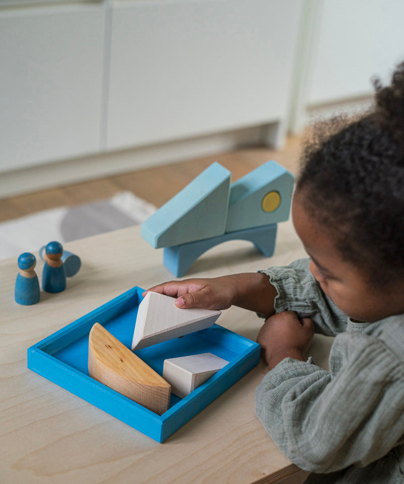 A child playing with the Grimm's blue boat Wooden Stacking Puzzle. The child is placing the blocks into the blue coloured wooden tray. These Grimm's toys are part of a wide range of wooden toys available here at Babipur. 