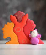Grimm's handcrafted wooden casa glora set with a small dwarf doll standing on top. Building blocks in warm red and orange colours
