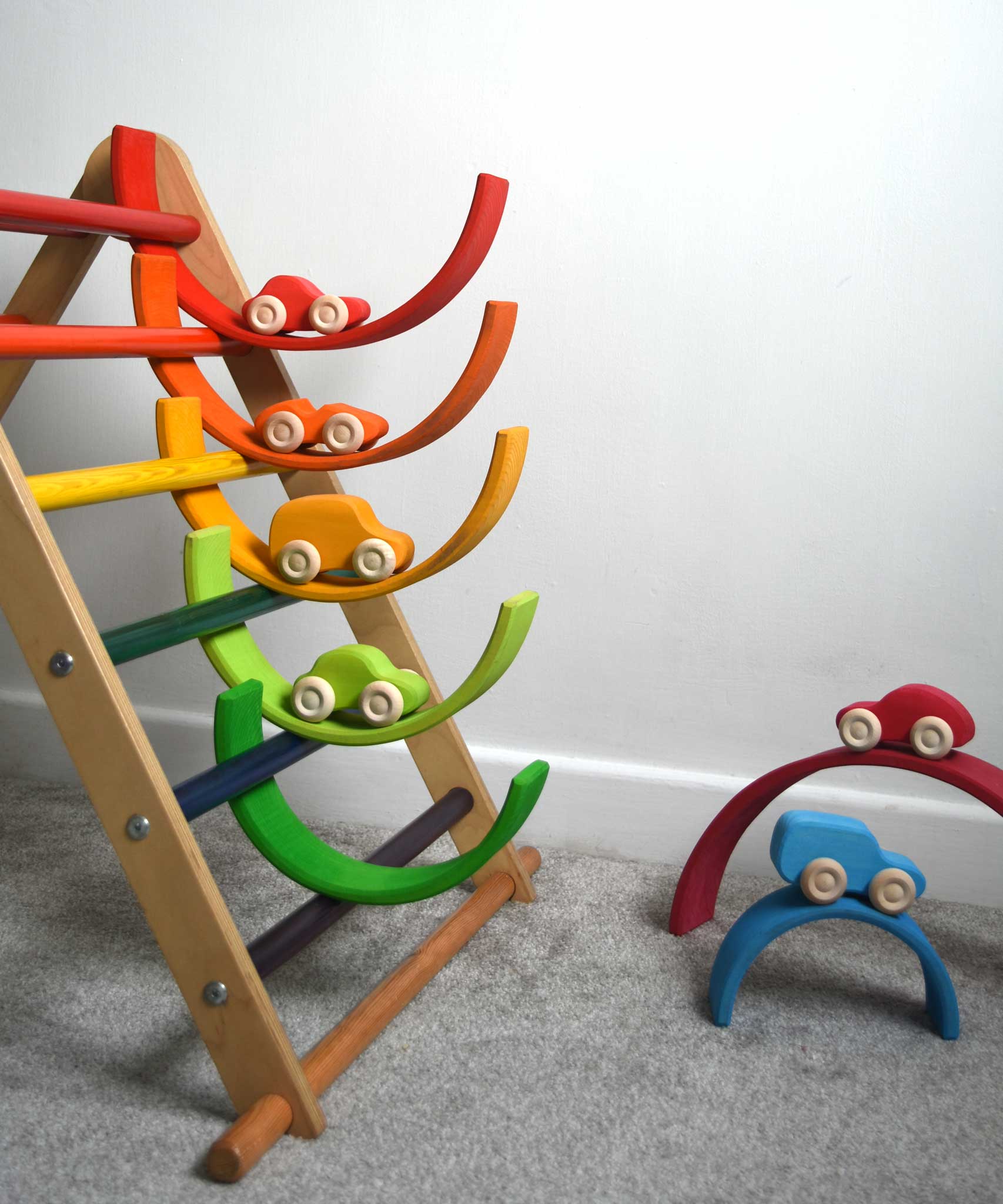 The cars from the Grimm's 6 Coloured Wooden Cars set placed on matching coloured pieces from the large Grimm's rainbow tunnel. Some pieces of the rainbow have been placed in the rungs of the Triclimb climbing frame. These wooden toy cars are part of a wide range of Grimm's wooden toys available here at Babipur.