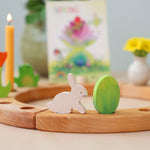 Grimm's Easter Decorative Figure Set featuring a white bunny rabbit a Batik Green coloured eggs. The figures are shown on a natural wooden celebration ring. Other decorative figures can be seen placed on the ring in the background. 