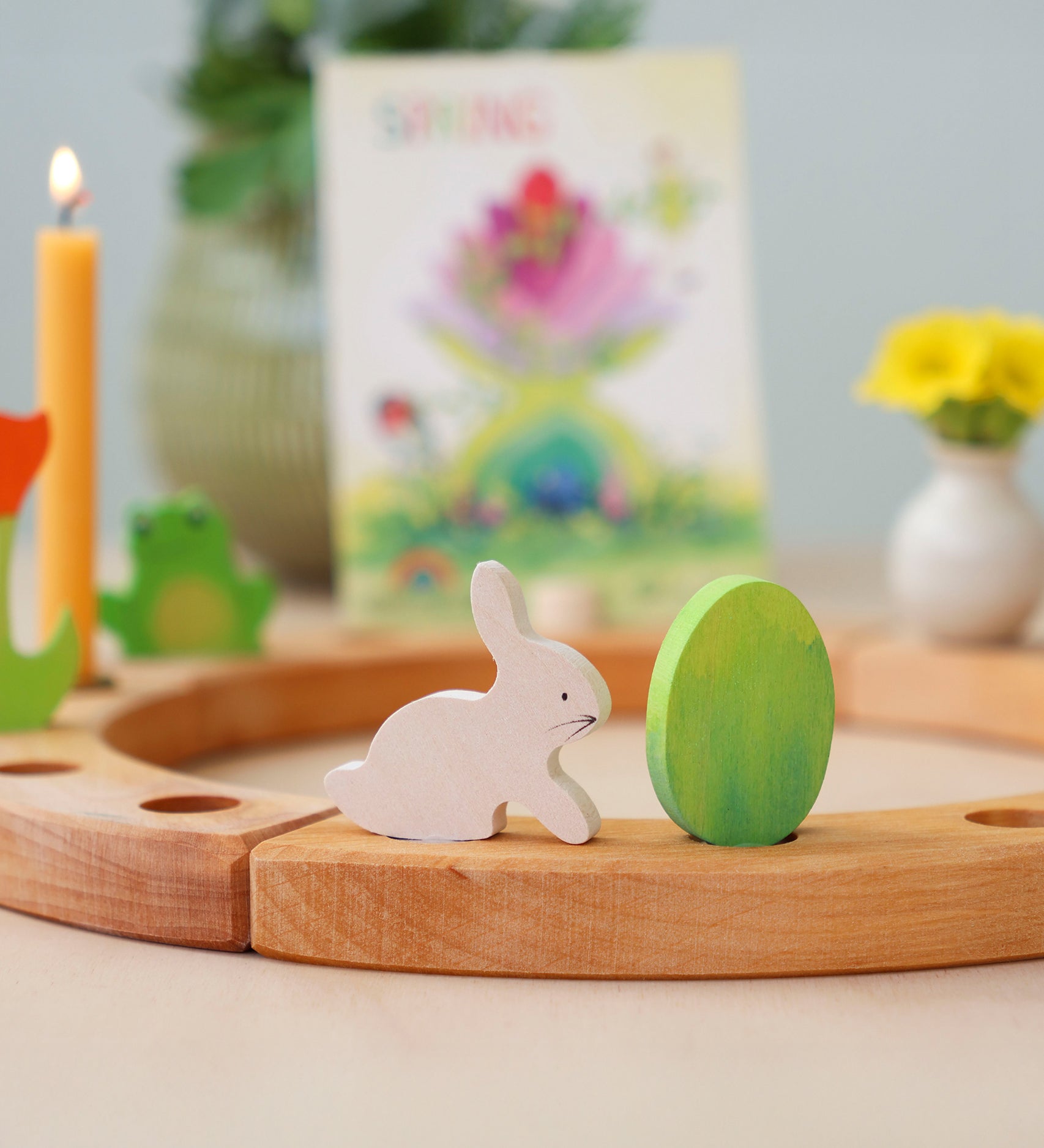 Grimm's Easter Decorative Figure Set featuring a white bunny rabbit a Batik Green coloured eggs. The figures are shown on a natural wooden celebration ring. Other decorative figures can be seen placed on the ring in the background. 