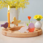 A Grimm's Easter Decorative Figure Set. The set includes a white bunny rabbit figure and a Batik Orange and purple coloured egg shown on a natural wooden celebration ring. Other figures have been paced on the ring can be seen in the background.  