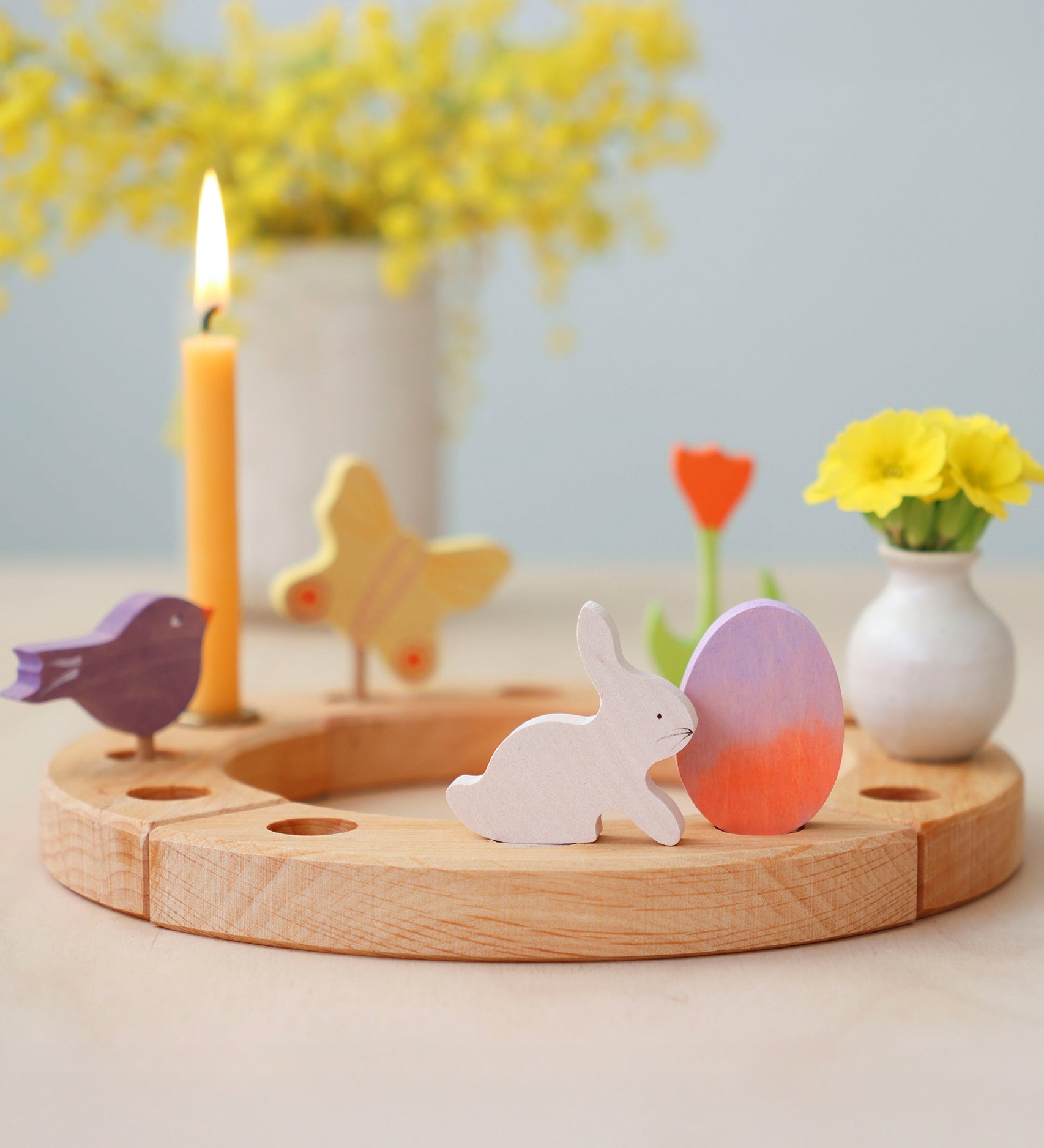 A Grimm's Easter Decorative Figure Set. The set includes a white bunny rabbit figure and a Batik Orange and purple coloured egg shown on a natural wooden celebration ring. Other figures have been paced on the ring can be seen in the background.  