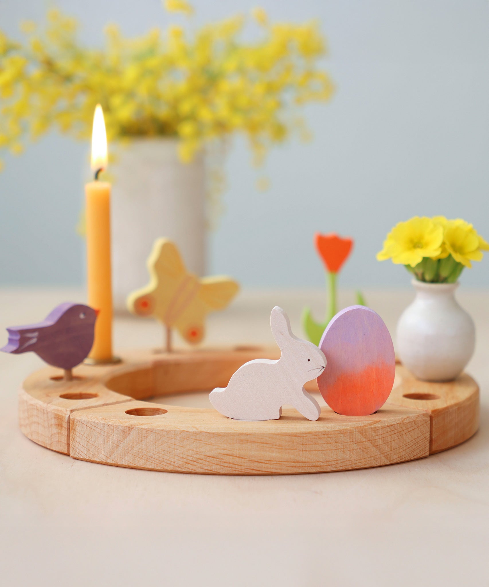A Grimm's Easter Decorative Figure Set. The set includes a white bunny rabbit figure and a Batik Orange and purple coloured egg shown on a natural wooden celebration ring. Other figures have been paced on the ring can be seen in the background.  