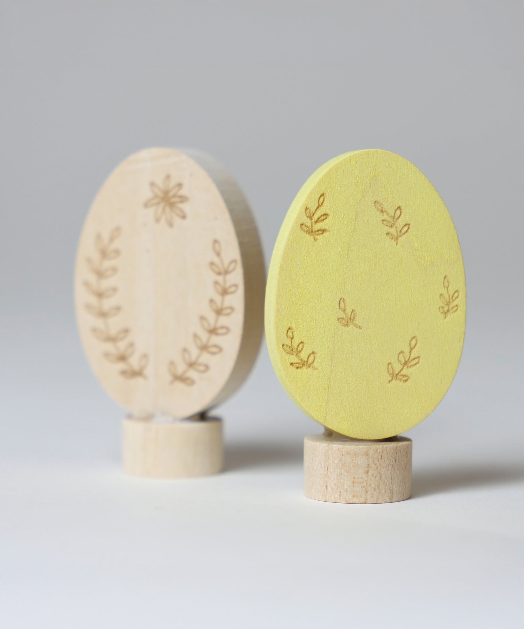 Grimm's Ornamental Duo Easter Eggs Decorative Figure Set. One egg is left natural withe floral themed outline details, the other painted in a light yellow with leaf outline details. 