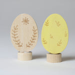 Grimm's Ornamental Duo Easter Eggs Decorative Figure Set. One egg is left natural withe floral themed outline details, the other painted in a light yellow with leaf outline details. 
