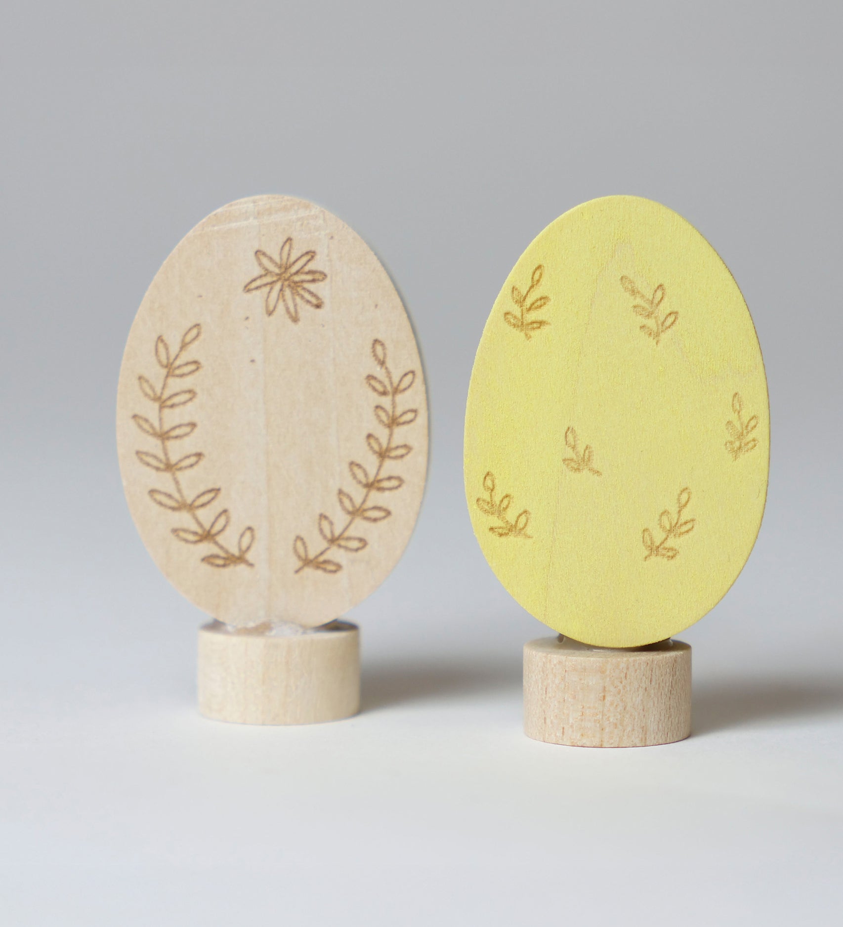 Grimm's Ornamental Duo Easter Eggs Decorative Figure Set. One egg is left natural withe floral themed outline details, the other painted in a light yellow with leaf outline details. 