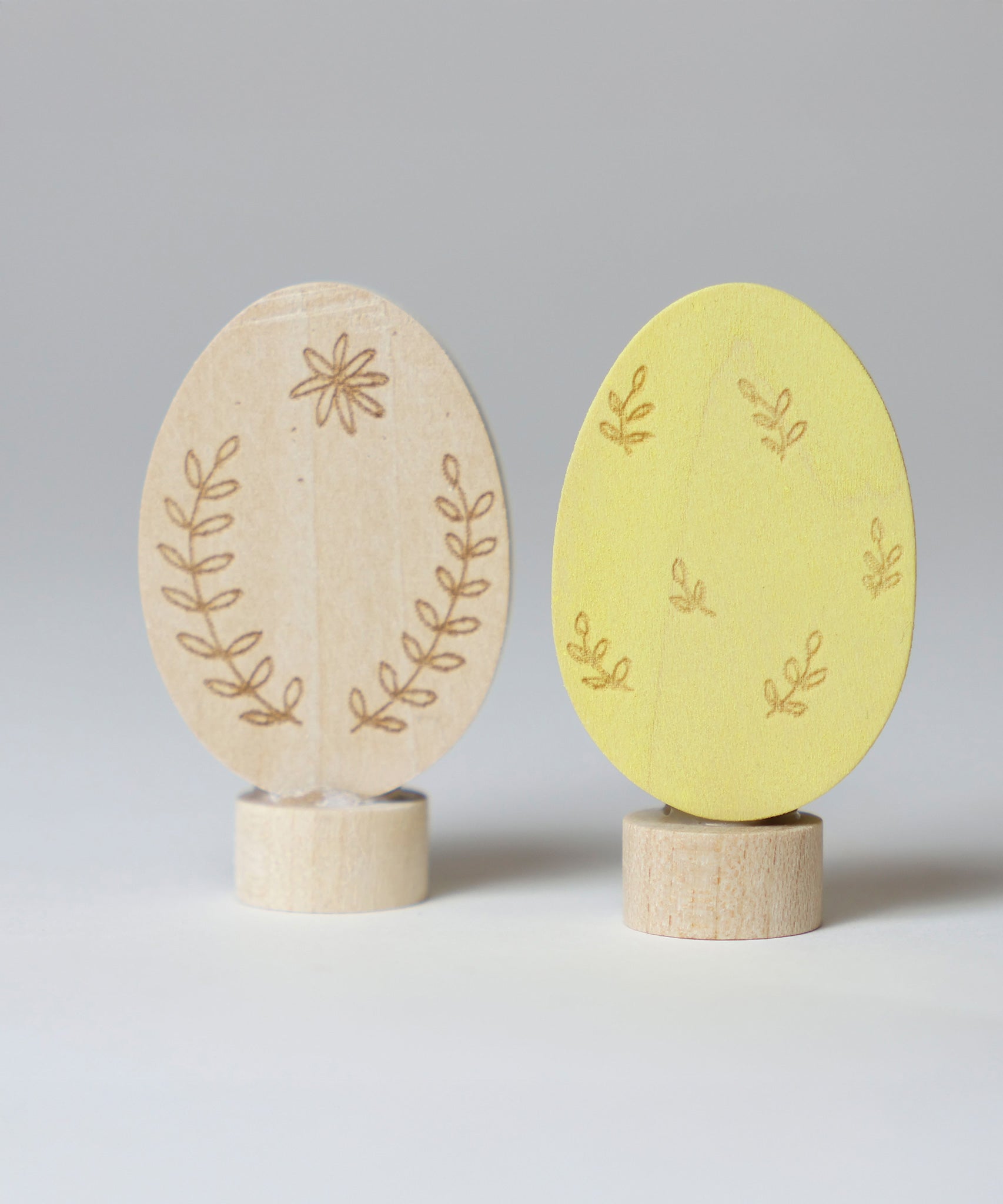 Grimm's Ornamental Duo Easter Eggs Decorative Figure Set. One egg is left natural withe floral themed outline details, the other painted in a light yellow with leaf outline details. 