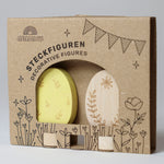 Grimm's Ornamental Duo Easter Eggs Decorative Figure Set. One egg is left natural withe floral themed outline details, the other painted in a light yellow with leaf outline details. The eggs are shown in their cardboard packaging sleeve. 