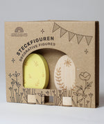 Grimm's Ornamental Duo Easter Eggs Decorative Figure Set. One egg is left natural withe floral themed outline details, the other painted in a light yellow with leaf outline details. The eggs are shown in their cardboard packaging sleeve. 