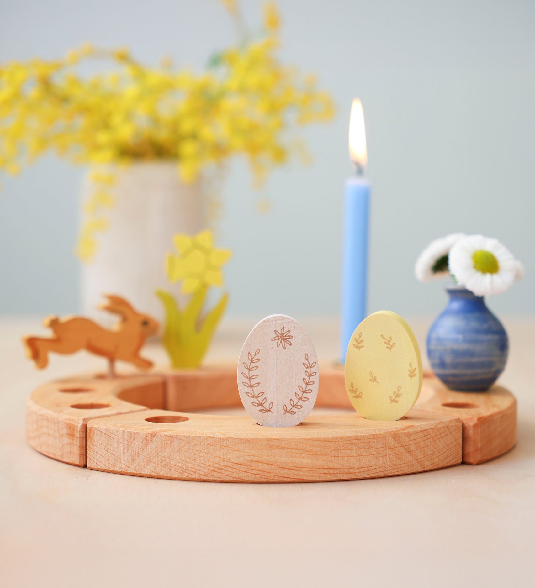 Grimm's Ornamental Duo Easter Eggs Decorative Figure Set. The eggs are placed on a natural wooden Grimm's celebration ring. Other decorative figures and a blue coloured candle can be seen in the background. 