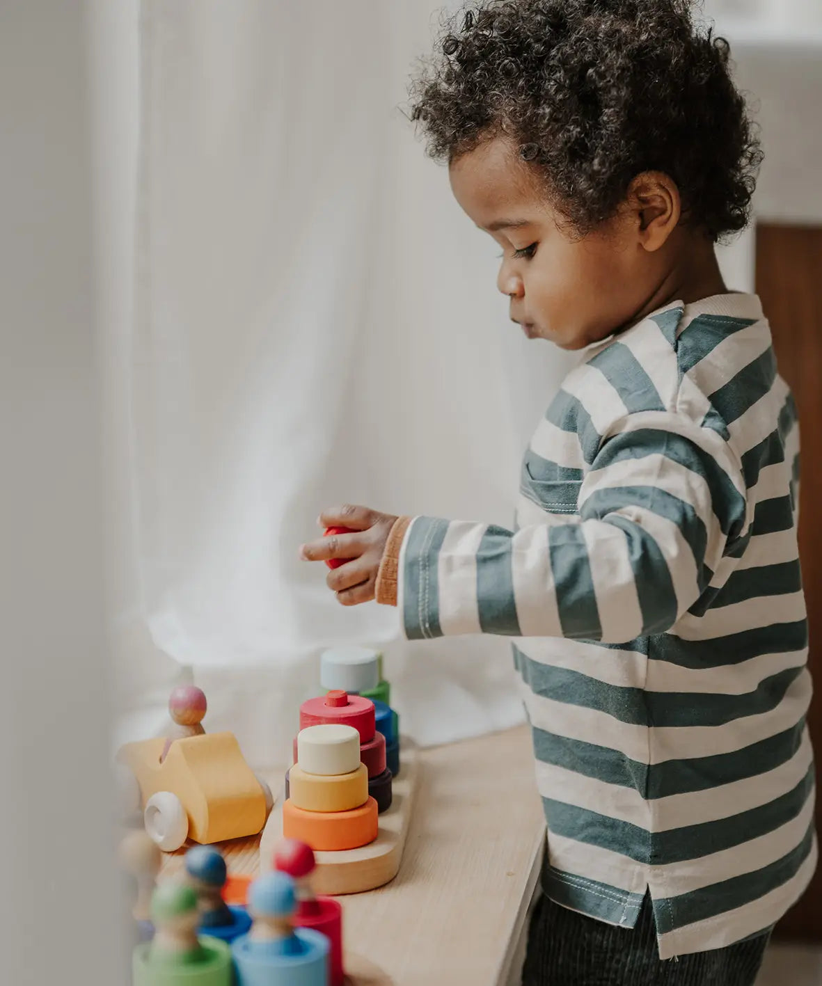 Child standing over a Grimms fabuto stacking set and holding a loose red disk