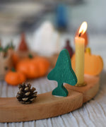 A Grimm's fir tree decorative figure placed on a natural wooden celebration ring. A lit candle and other Autumn themed decorations can be seen in the background. The decorative figures are part of a wide range of Waldorf inspired figures and celebration rings available here at Babipur. 