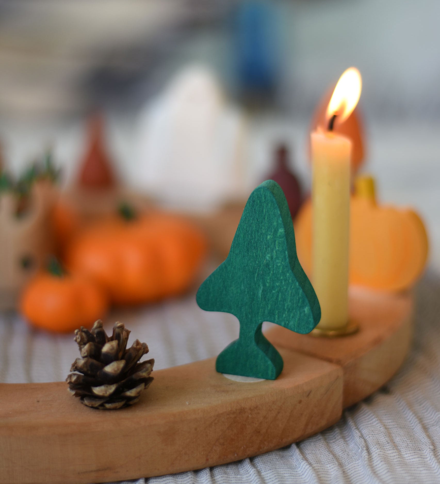 A Grimm's fir tree decorative figure placed on a natural wooden celebration ring. A lit candle and other Autumn themed decorations can be seen in the background. The decorative figures are part of a wide range of Waldorf inspired figures and celebration rings available here at Babipur. 